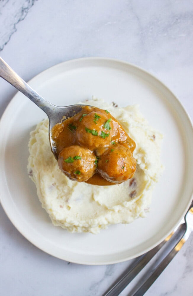 A spoonful of meatballs in brown gravy is held over a plate of mashed potatoes on a white plate with a fork and knife beside it.