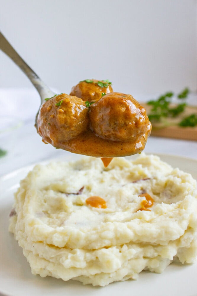 A spoon holding three meatballs in brown gravy is positioned above a plate of mashed potatoes.