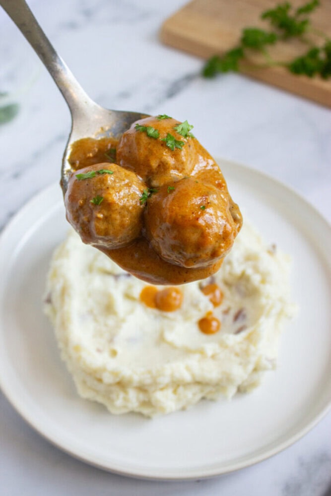 A spoonful of meatballs in brown gravy held above a plate of mashed potatoes.