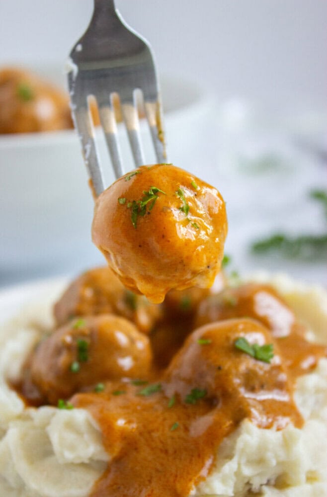 A fork holds a saucy meatball above a plate of mashed potatoes topped with several meatballs and garnished with chopped herbs.