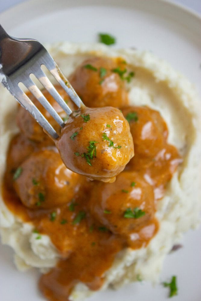 A fork holds a meatball covered in brown gravy above a plate of mashed potatoes topped with more meatballs and sauce, garnished with chopped herbs.