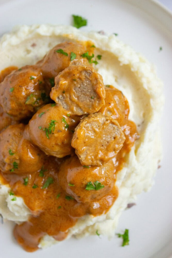 A serving of meatballs in creamy brown sauce on a bed of mashed potatoes, garnished with chopped parsley.