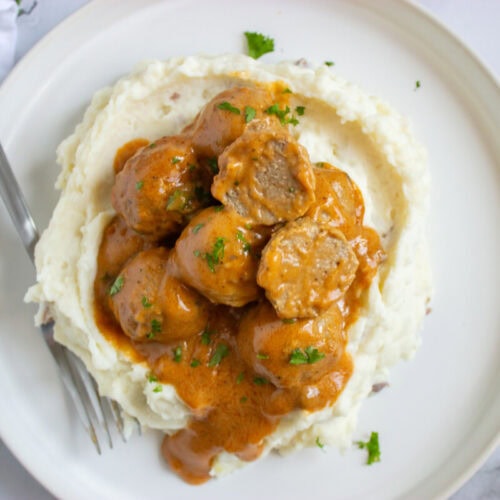 A plate of mashed potatoes topped with meatballs in brown gravy, garnished with chopped herbs, with a fork on the side.