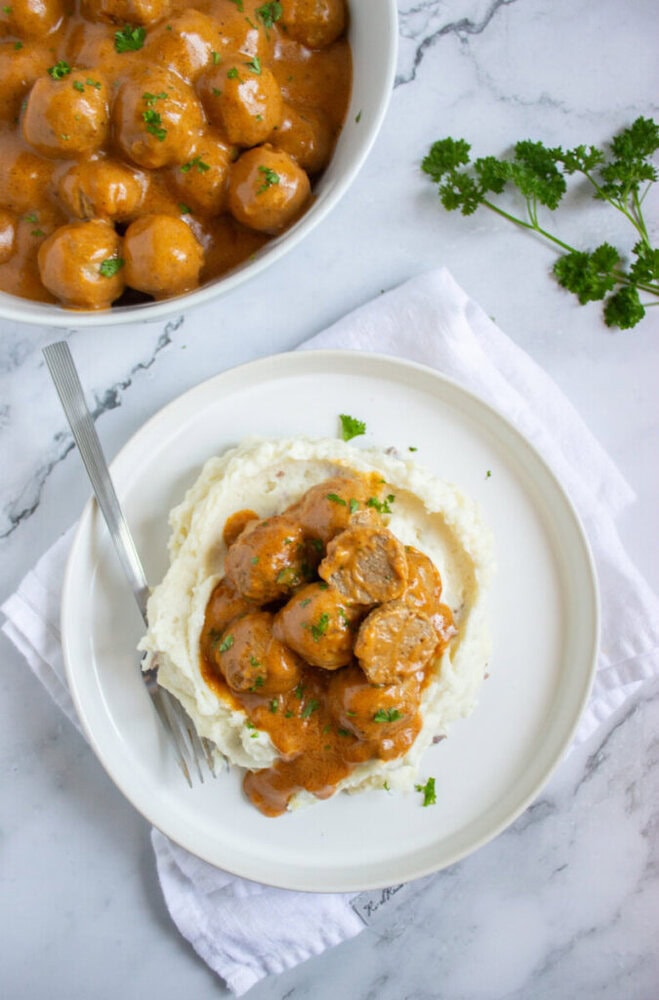 Meatballs in creamy brown sauce served over mashed potatoes on a white plate, garnished with parsley, with a fork and a bowl of extra meatballs nearby.