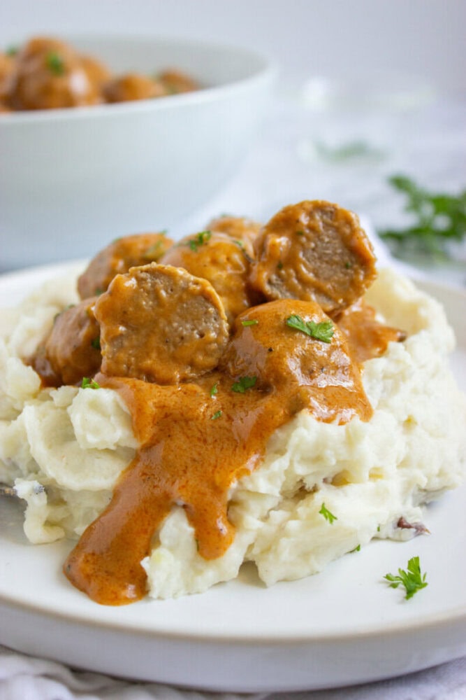 A plate of mashed potatoes topped with meatballs and brown gravy, garnished with chopped parsley.