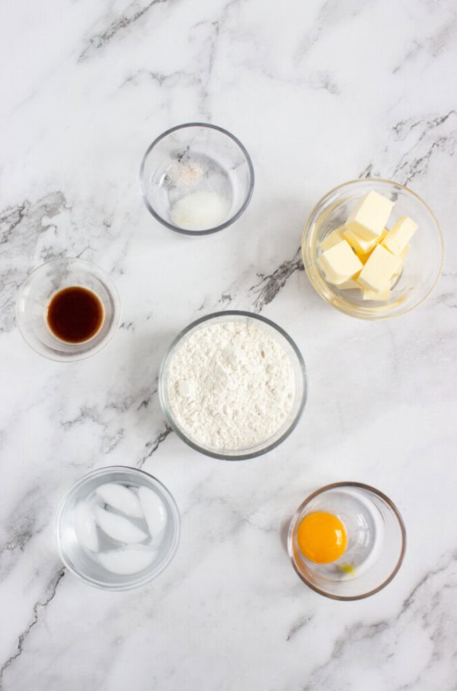 Six glass bowls containing butter, flour, an egg, vanilla extract, salt and sugar, and ice water arranged on a white marble surface.