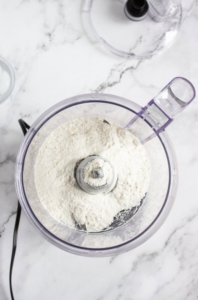 A food processor bowl filled with flour sits on a marble countertop, with the appliance lid and cord visible nearby.