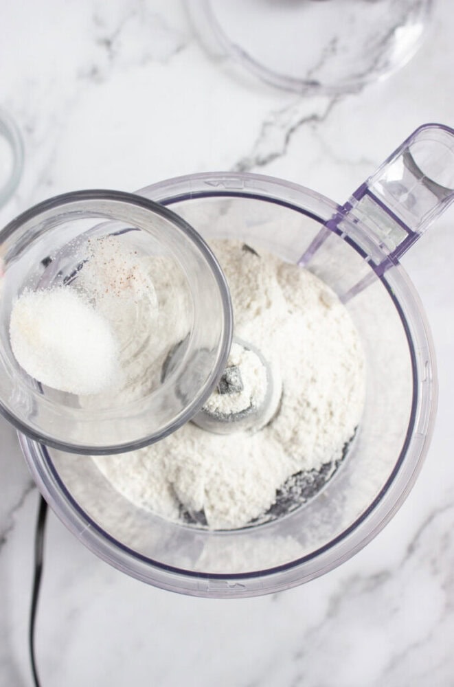 A clear bowl of flour in a food processor with a small container of salt and seasoning being added, on a marble countertop.