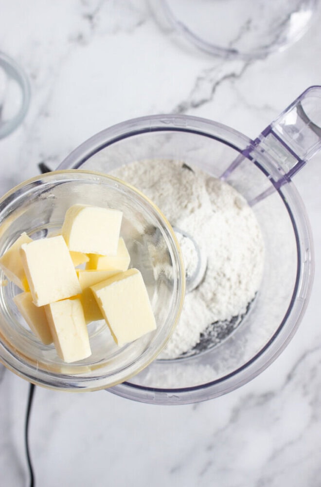 Cubes of butter in a glass bowl are about to be added to a food processor containing flour on a marble countertop.