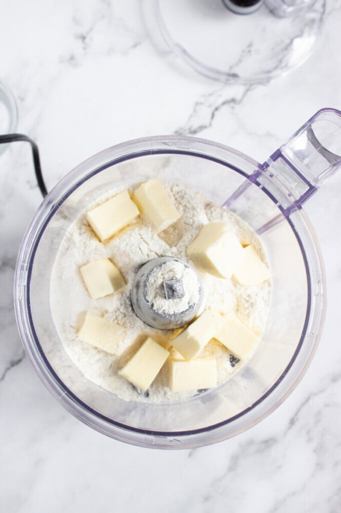 A food processor bowl with flour, sugar, and cubes of butter inside, ready for mixing, on a marble countertop.
