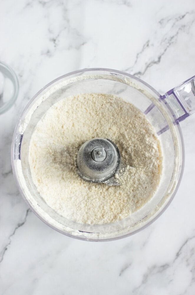 A food processor filled with finely ground flour is placed on a white marble countertop.