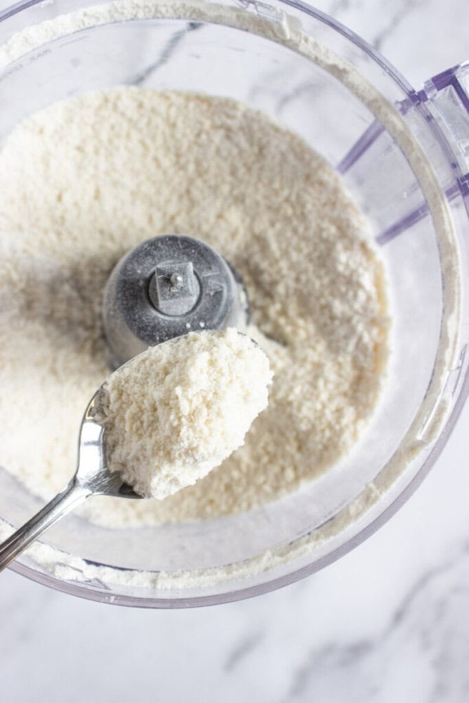 A spoon holds finely ground almond flour above a food processor filled with more almond flour, on a white surface.