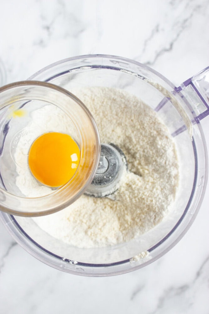 A glass bowl with a raw egg is being poured into a food processor containing flour on a marble countertop.