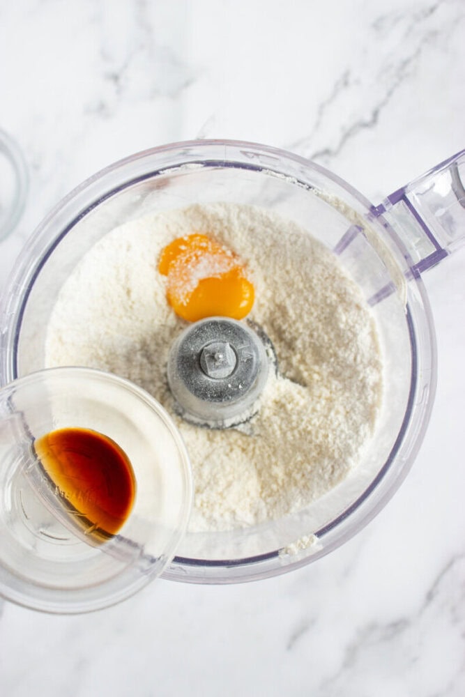 A food processor bowl with flour, an egg yolk, and salt, while a hand adds vanilla extract from a small glass cup.
