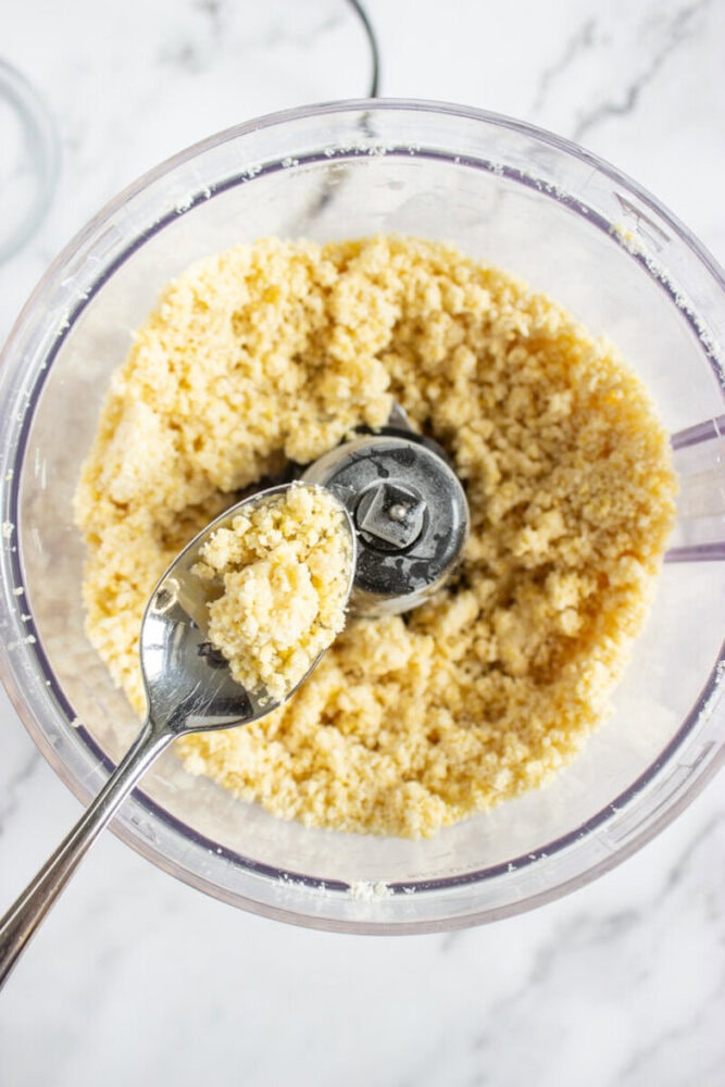 Food processor bowl with crumbly dough mixture inside, a spoon holding some of the mixture is positioned above the blades.