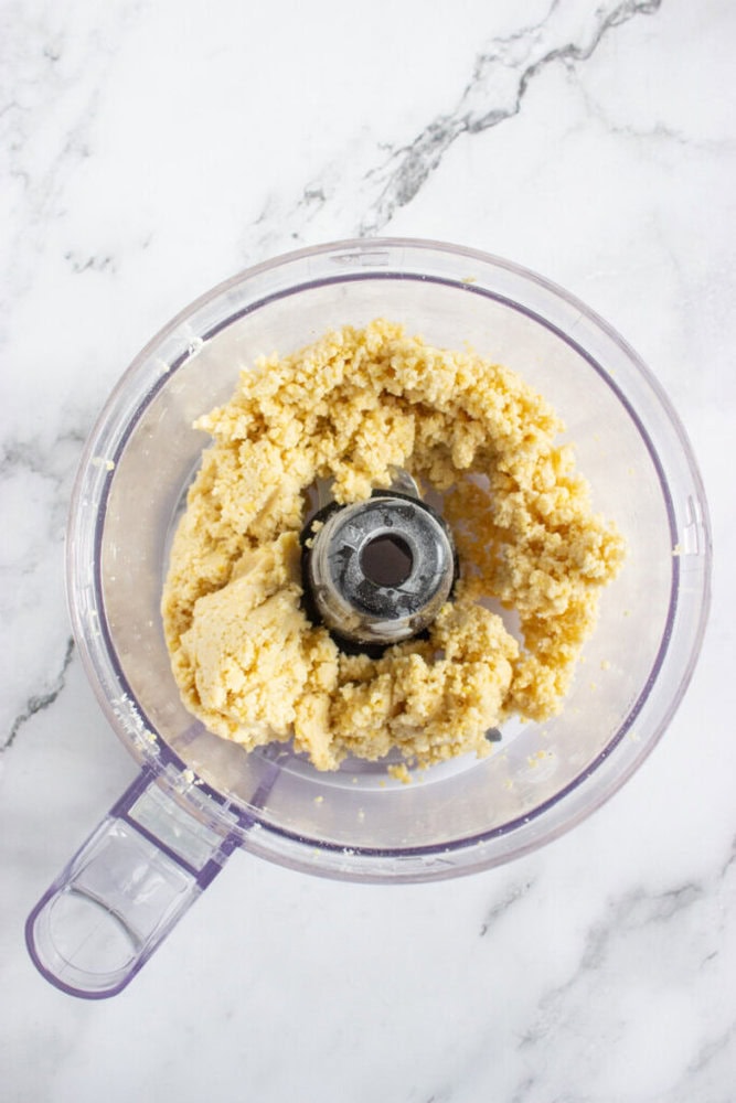 A food processor bowl containing a coarse, pale yellow dough mixture on a white marble surface.