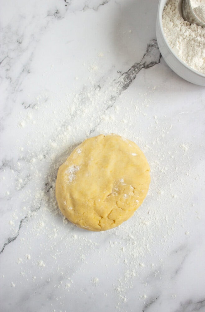 A round ball of dough rests on a floured marble surface, with a bowl of flour and a metal scoop partially visible in the top right corner.