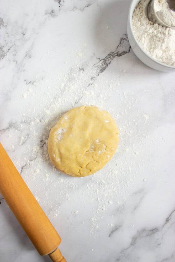 A round disk of dough on a floured marble surface, with a rolling pin and a bowl of flour nearby.