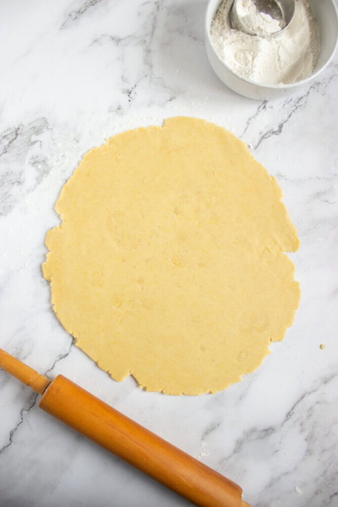 A rolled-out sheet of dough on a marble surface with a rolling pin below and a bowl of flour with a scoop above.