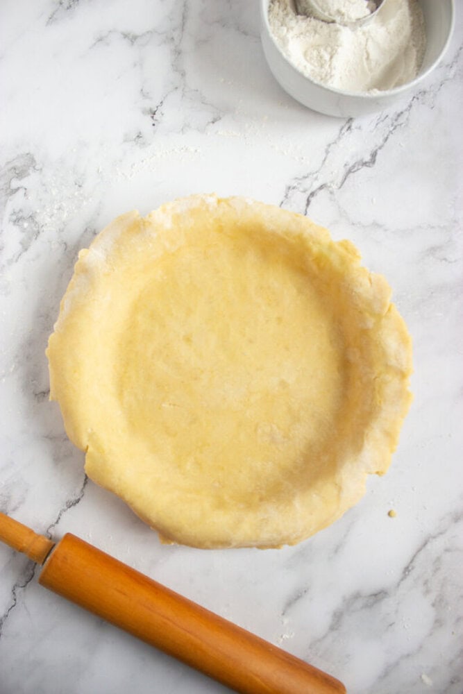 Unbaked pie crust in a pie dish on a marble surface, with a rolling pin and a bowl of flour nearby.