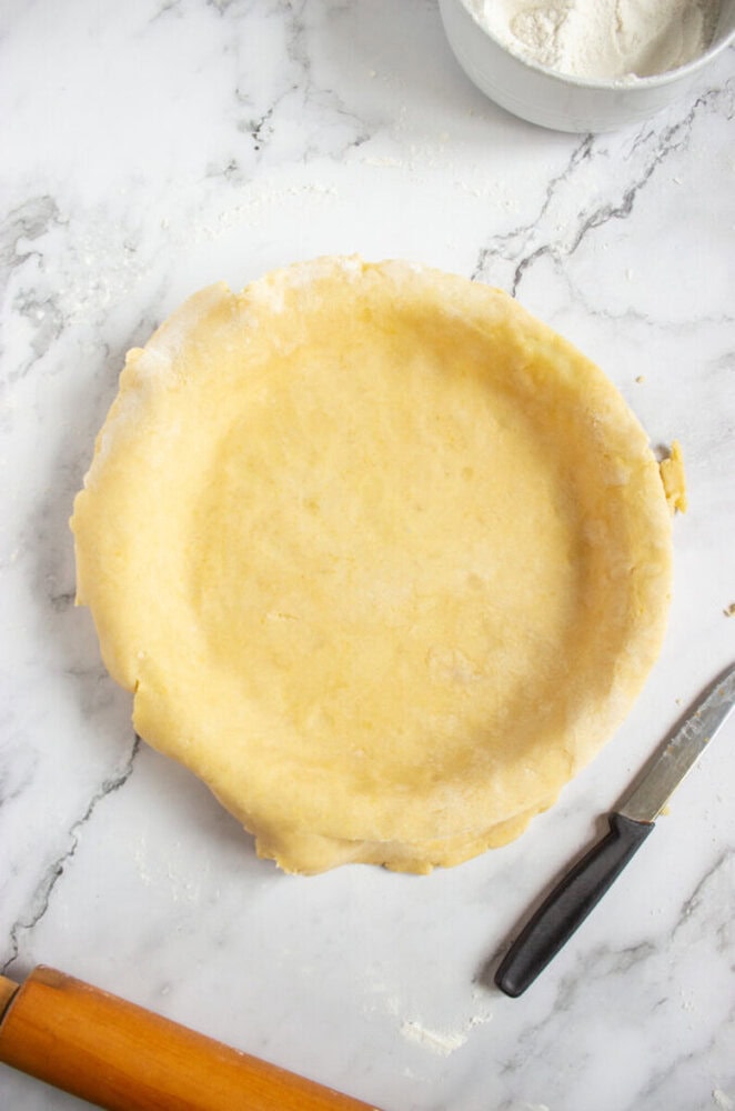 Unbaked pie crust in a round dish on a marble surface, with a rolling pin, knife, and a bowl of flour nearby.