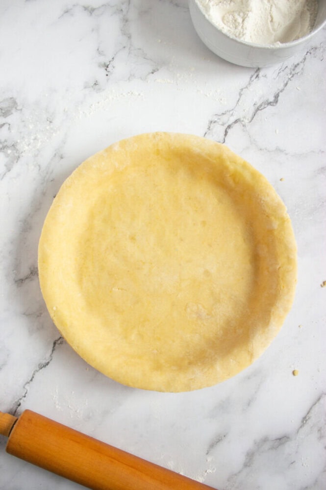 Unbaked pie crust in a round dish on a marble surface, with a rolling pin and a bowl of flour nearby.
