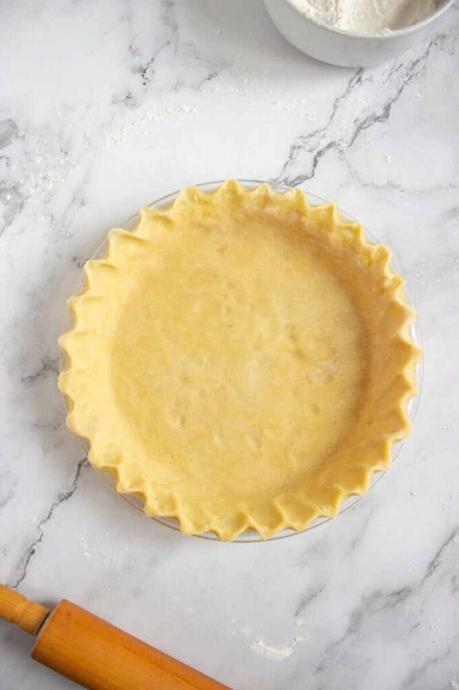 Unbaked pie crust in a glass pie dish on a marble surface, with a rolling pin and a bowl of flour nearby.