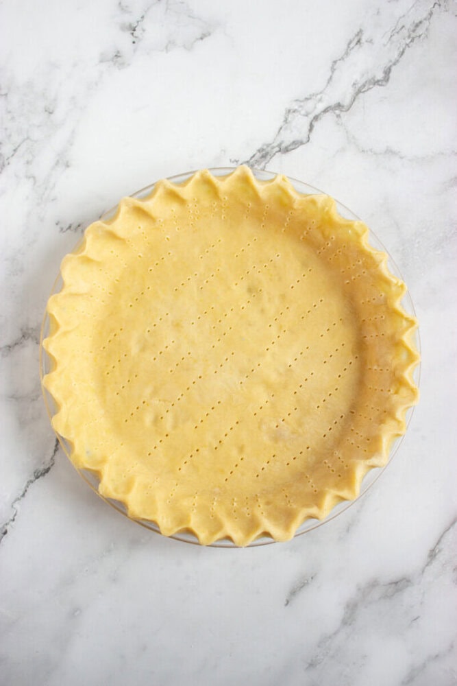 Unbaked pie crust with crimped edges and fork pricks sits in a glass pie dish on a marble surface.