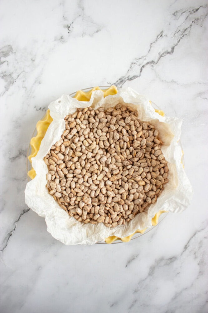 A pie dish lined with parchment paper is filled with dried beans, used as pie weights, on a marble surface.