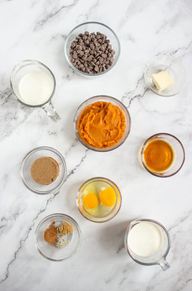 Overhead view of various baking ingredients in glass bowls and cups on a marble surface, including eggs, milk, pumpkin puree, chocolate chips, brown sugar, spices, butter, and vanilla extract.