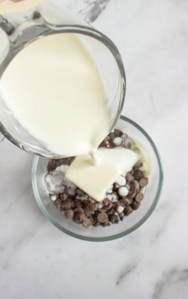 A glass measuring cup pours cream over chocolate chips and a pat of butter in a clear glass bowl on a marble surface.