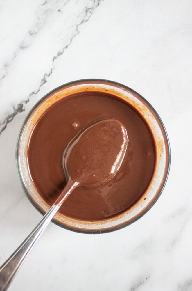 A spoon rests in a bowl filled with smooth, glossy melted chocolate on a white marble surface.
