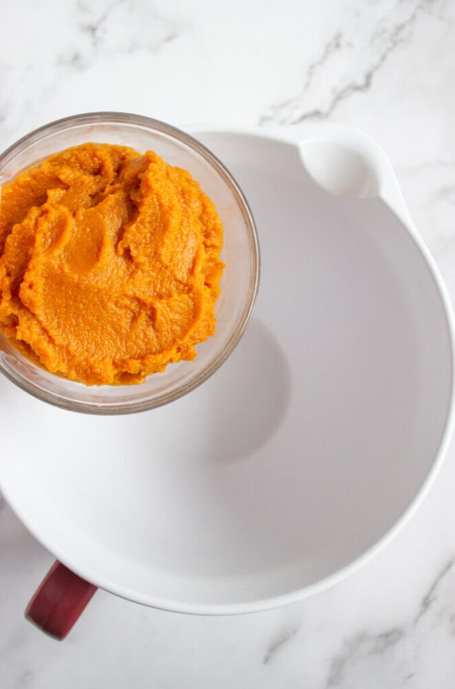 A glass bowl of orange pumpkin puree sits on the edge of a large, empty white mixing bowl on a marble countertop.