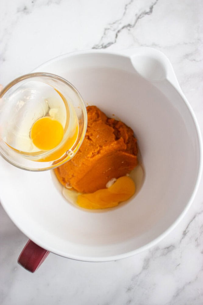 A hand holds a bowl with an egg above a mixing bowl containing pumpkin puree and another cracked egg on a marble countertop.