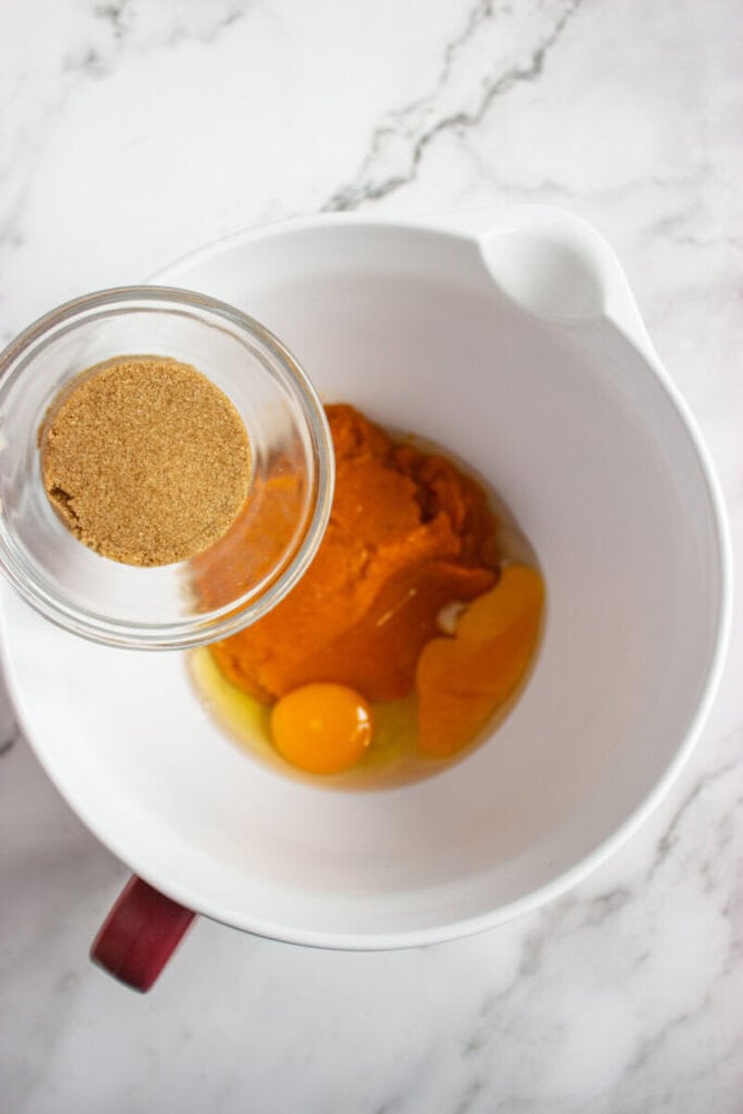 A white mixing bowl containing pumpkin puree and an egg, with a small glass bowl of brown sugar being held above it on a marble countertop.