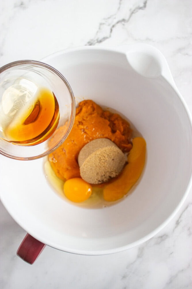 A mixing bowl with pumpkin puree, brown sugar, and two eggs, with a small bowl of vanilla extract being poured in, set on a white marble surface.
