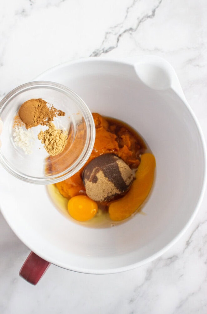 A white mixing bowl with eggs, brown sugar, and pumpkin puree. A hand holds a small bowl of ground spices above the main bowl on a marble countertop.
