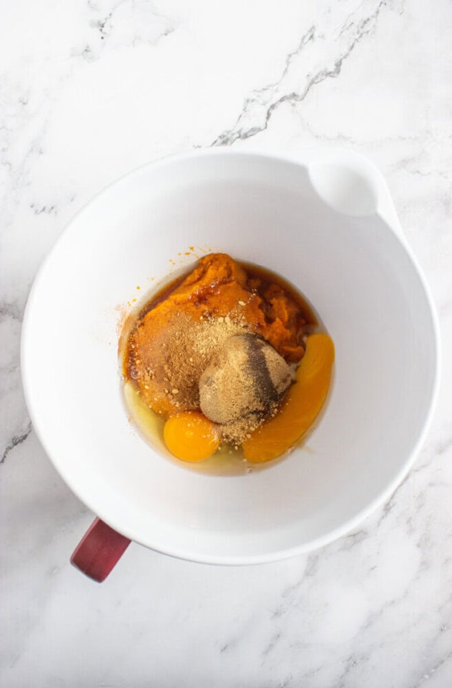 A white mixing bowl containing eggs, pumpkin puree, brown sugar, and spices on a marble countertop.