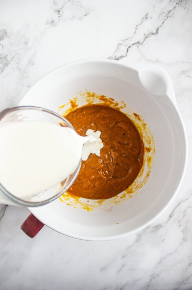A white bowl contains an orange batter, with milk being poured in from a glass measuring cup on a marble countertop.