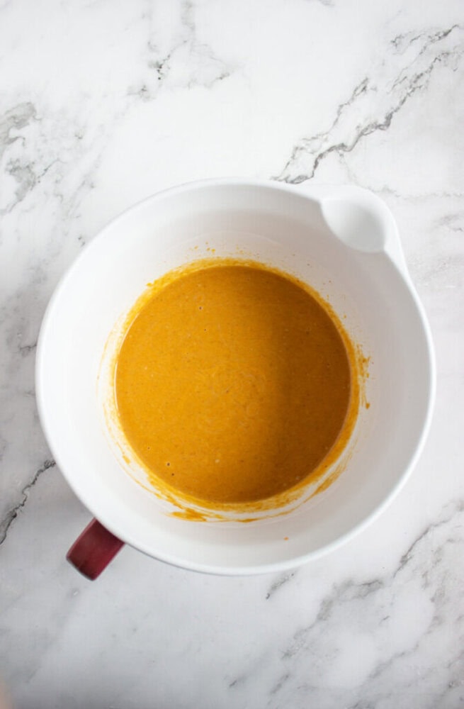 A white mixing bowl with orange batter inside sits on a white marble countertop.
