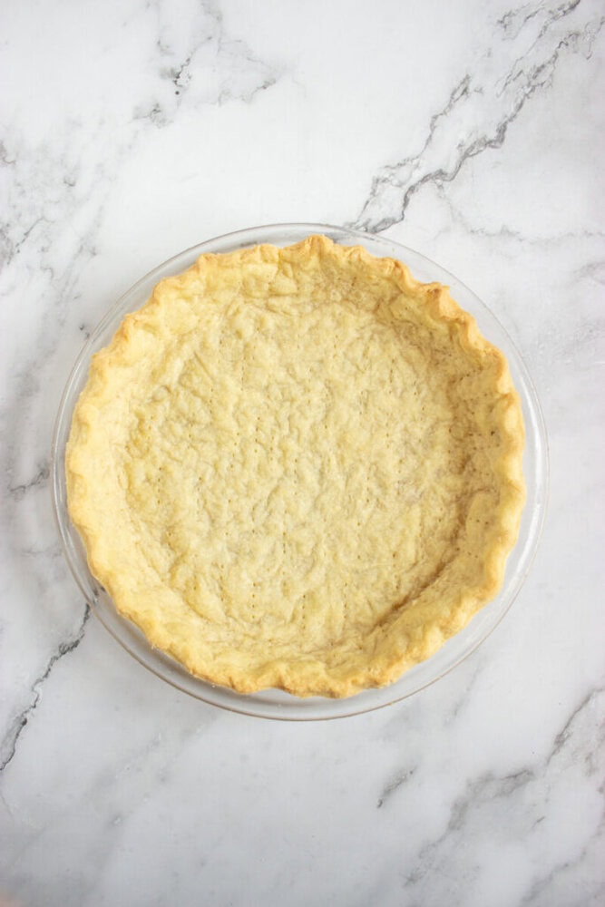 A baked, empty pie crust sits in a clear glass pie dish on a white marble surface.