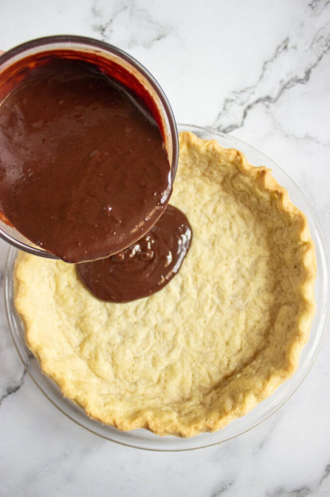 Chocolate filling is being poured from a bowl into an unbaked pie crust on a marble countertop.