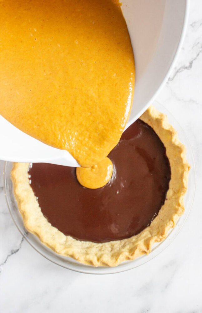 Pumpkin pie filling is being poured from a white bowl into a pie crust that already contains a layer of chocolate filling.