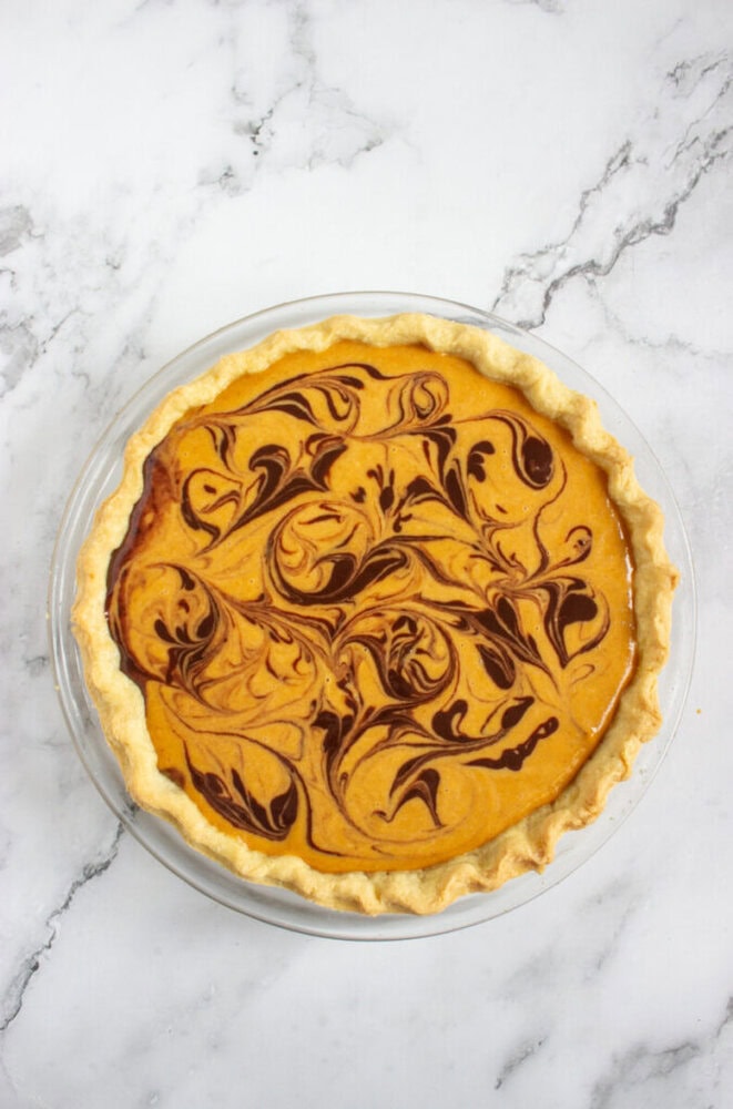 A pumpkin pie in a glass pie dish with a chocolate swirl pattern on top, set on a white marble surface.