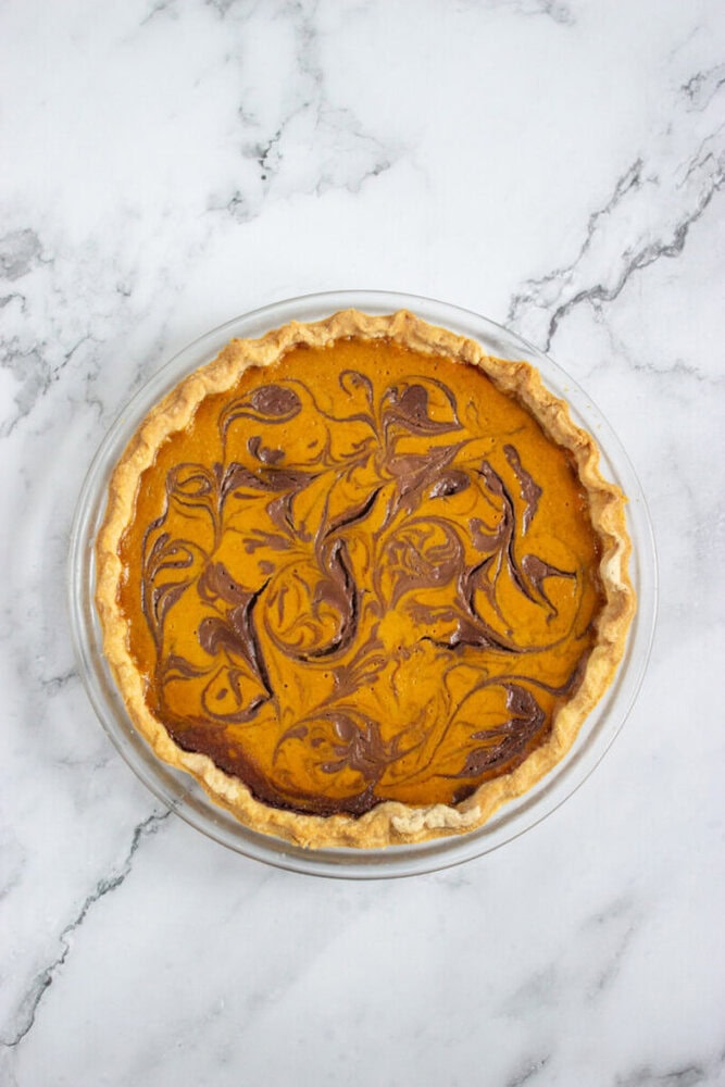 A pumpkin pie with a marbled chocolate swirl pattern on top, set in a clear glass pie dish on a white marble surface.