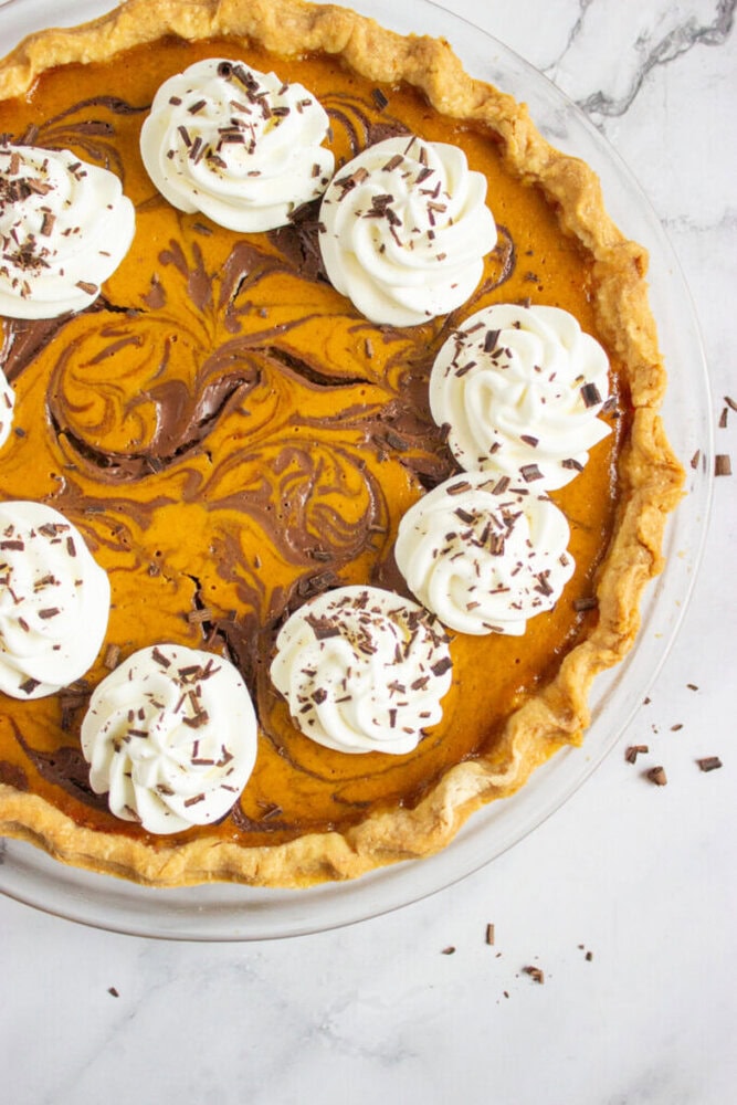 Pumpkin pie with a chocolate swirl design, topped with dollops of whipped cream and chocolate shavings, on a marble surface.