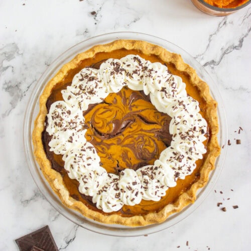 A pumpkin and chocolate swirl pie topped with whipped cream and chocolate shavings, placed on a marble surface next to a bowl of pumpkin puree and pieces of chocolate.