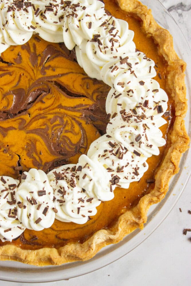 A pumpkin pie with a marbled chocolate swirl, topped with a ring of piped whipped cream and chocolate shavings on a golden crust.