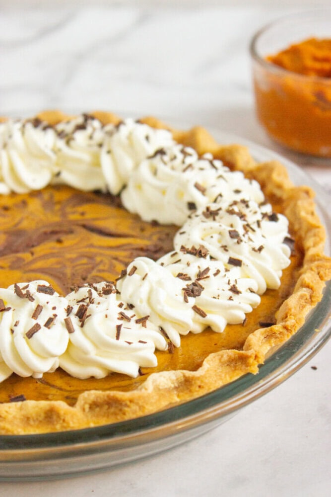 A pie with a swirled filling, topped with whipped cream and chocolate shavings, sits in a glass dish on a marble surface; a bowl of puree is in the background.