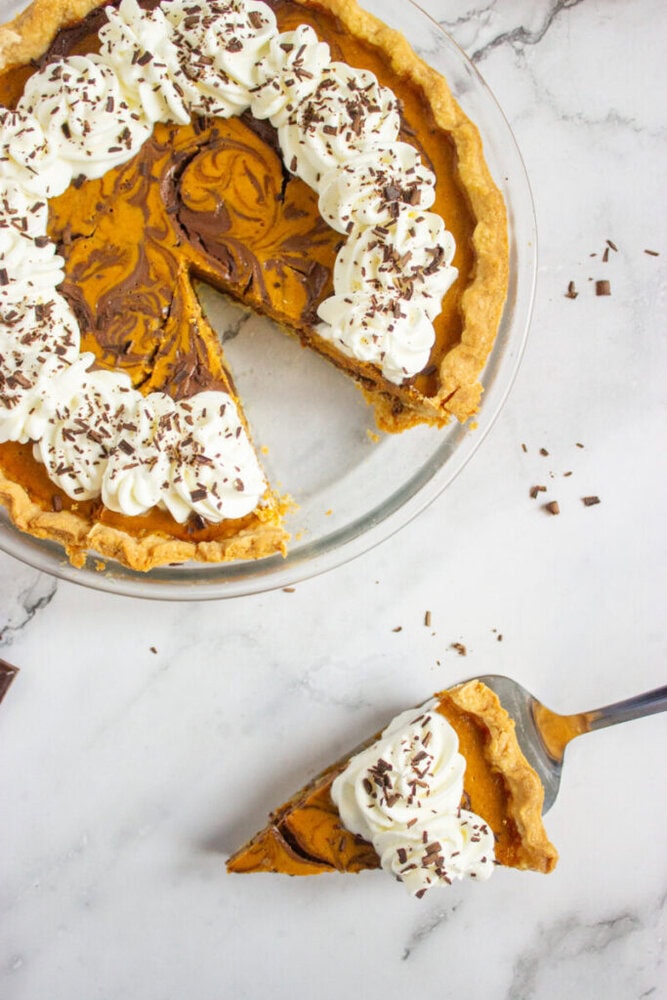 A marbled pie with a slice removed sits in a glass dish. The pie is topped with whipped cream and chocolate shavings, and one slice is being served on a spatula.